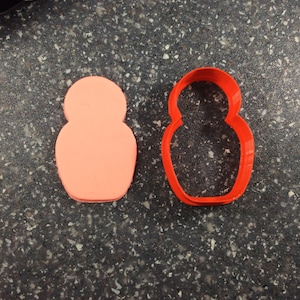 May include: A red plastic cookie cutter shaped like a traditional Russian nesting doll, next to a pink cookie dough cutout in the same shape.