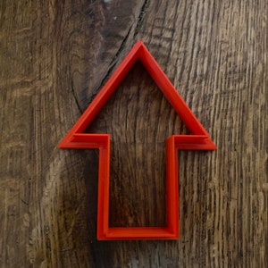 May include: A red plastic arrow-shaped cookie cutter. The cutter is on a wooden surface.