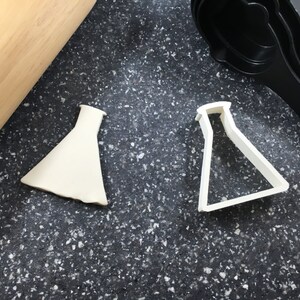 May include: Two white plastic cookie cutters shaped like beakers on a black speckled countertop. One cutter is a flat shape and the other is a 3D shape.
