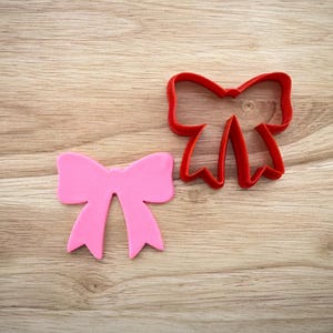 May include: A pink bow-shaped cookie cutter next to a red bow-shaped cookie cutter. The pink bow is a solid shape, while the red cutter is an outline. Both are on a light brown wooden surface.