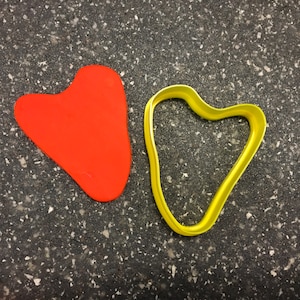 May include: A red heart-shaped cookie cutter and a yellow heart-shaped cookie cutter on a gray speckled surface.
