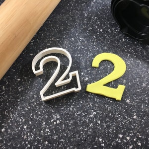 May include: Two cookie cutters in the shape of the number 2. One cutter is white plastic and the other is yellow.