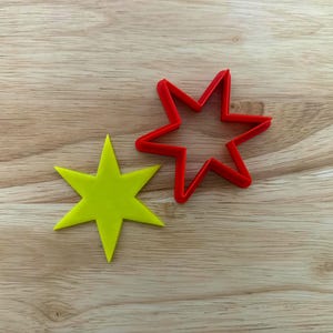 May include: A bright yellow star-shaped object and a red star-shaped cookie cutter sit on a wooden surface. The star has six points. The cookie cutter is made of red plastic. The wooden surface has a light brown color.