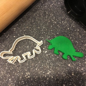 May include: A white plastic dinosaur cookie cutter and a green dinosaur-shaped cookie. A wooden rolling pin and a black kitchen tool are also in the image. The cookie cutter is in the shape of a Stegosaurus.