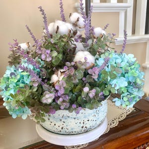 May include: A decorative floral arrangement in a speckled blue and white ceramic pot. The arrangement includes cotton bolls, lavender sprigs, green and purple foliage, and blue-green hydrangeas. The pot sits on a white saucer.