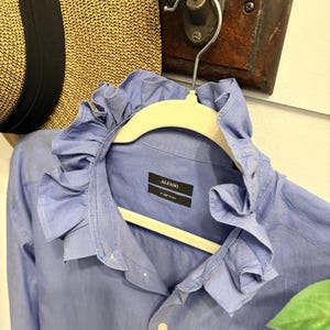 May include: A light blue button-down shirt featuring a prominent ruffled collar. The interior neck label displays the brand name Alfani and the word Pinpoint. The shirt is hung on a white hanger, showcasing long sleeves and a front button placket.