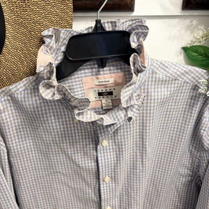 May include: A long-sleeve button-down shirt featuring a light blue and tan gingham check pattern. The shirt has a high, ruffled collar and a label inside the neck that reads Joseph Abboud, Non-Iron, and size XL. The fabric is 100% cotton.