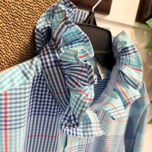 May include: A long-sleeved button-down shirt featuring a light blue, dark blue, and white plaid pattern. The shirt has a prominent ruffled collar made from the same plaid fabric, a front button placket, and a single visible light-colored button.