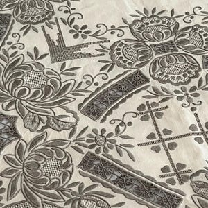 May include: Close-up of a cream-colored textile with intricate gray embroidery. The design features floral patterns, including large stylized flowers, along with geometric and scrollwork details. The embroidery is dense and detailed.