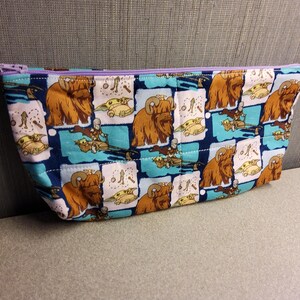 May include: A blue and white zippered pouch with a repeating pattern of brown furry creatures and small green characters. The pouch is made of fabric and has a purple zipper.