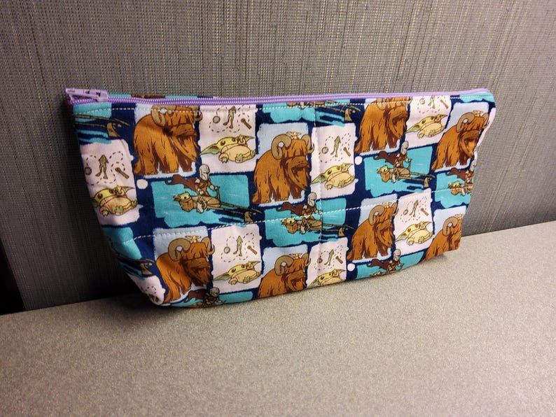 May include: A blue and white zippered pouch with a repeating pattern of brown furry creatures and small green characters. The pouch is made of fabric and has a purple zipper.