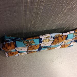 May include: A long, rectangular, blue, pink, and brown fabric pillow with a cartoon pattern. The pillow is lying on a light brown surface.