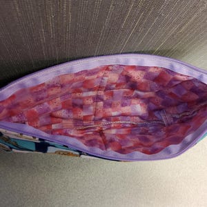 May include: A purple zippered pouch with a pink and purple checkered lining. The pouch is made of fabric and has a blue and white pattern on the outside.