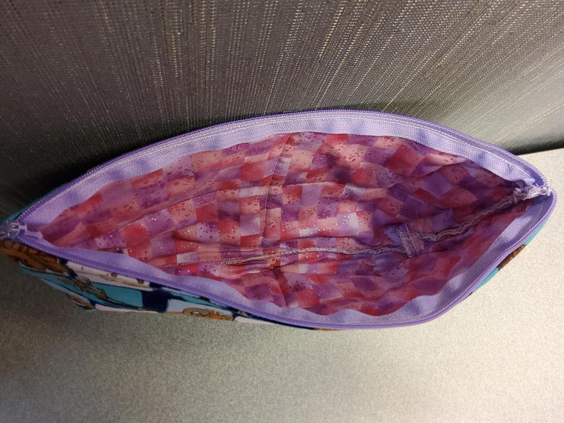 May include: A purple zippered pouch with a pink and purple checkered lining. The pouch is made of fabric and has a blue and white pattern on the outside.