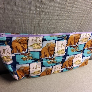 May include: A blue and white zippered pouch with a repeating pattern of brown furry creatures and green characters. The pouch has a purple zipper.