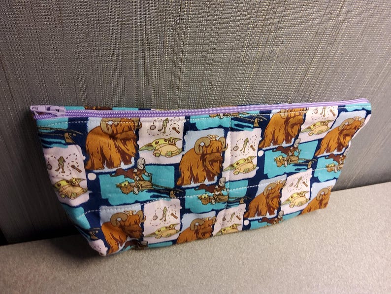 May include: A blue and white zippered pouch with a repeating pattern of brown furry creatures and green characters. The pouch has a purple zipper.