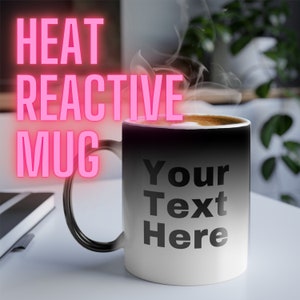 May include: A black heat reactive mug with the text "Your Text Here" printed on the front. The mug is filled with a hot beverage and steam is rising from the top.