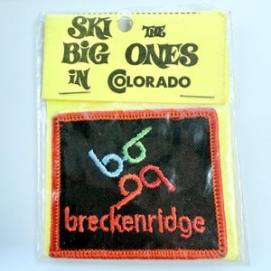 May include: A square embroidered patch with a black background and a red border. The patch features light blue and green abstract shapes above the orange text 'breckenridge'. It is attached to a yellow card printed with 'SKI THE BIG ONES IN COLORADO'.