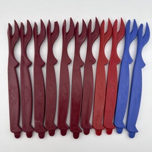 May include: A collection of plastic seafood shellers in various colors. The shellers are primarily burgundy, with a few red and blue ones. Each sheller has a long handle and a forked end for easy seafood removal.