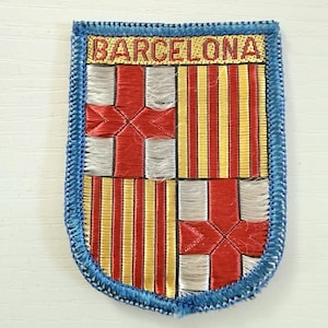 May include: A rectangular embroidered patch featuring the Barcelona coat of arms with a red cross on a white background and vertical yellow and red stripes. The word BARCELONA is stitched in red at the top, all bordered by a blue thread edge.
