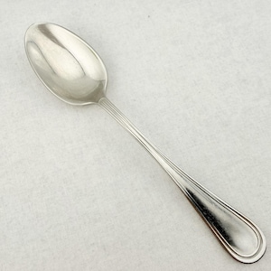 May include: A polished silver-toned metal spoon featuring a smooth, oval-shaped bowl and a slender handle. The handle is decorated with three parallel embossed lines that run along its length, terminating in a rounded, slightly flared end.