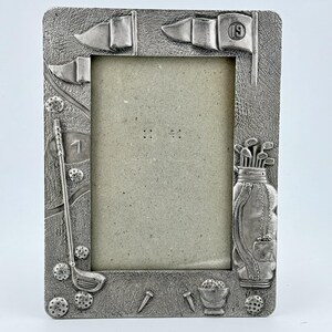 May include: A rectangular silver-colored picture frame with golf-themed embellishments. The frame features golf balls, flags, a golf bag, and a golf club. The flag on the top right has the number 19. The frame is designed to hold a photograph.