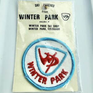 May include: A circular embroidered patch featuring a white background with a light blue border. The center displays a red stylized logo inside a triangle, with the red text WINTER PARK curved below. It is packaged in a clear plastic sleeve with a header card reading SKI PATCHES from WINTER PARK, AVAILABLE AT WINTER PARK SKI SHOP, WINTER PARK, COLORADO.