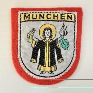 May include: A shield-shaped embroidered patch with a red felt border. The center features a silver background with the text MUNCHEN in black. A central figure with a yellow halo wears a black robe and holds a beer mug and a radish.