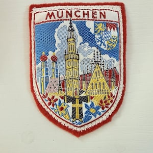 May include: A shield-shaped embroidered patch featuring the text MÜNCHEN at the top. The design depicts a city skyline with a tall central tower, two domed towers, and a coat of arms in the upper right corner, all bordered by a thick red edge.