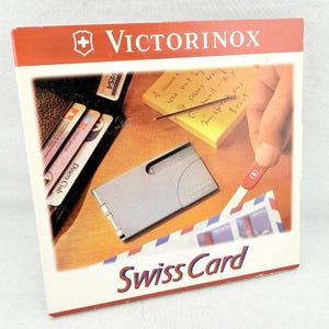 May include: A square product packaging box for a Victorinox Swiss Card. The box features a graphic showing a silver multi-tool card, a black wallet with credit cards, a yellow sticky note with handwriting, and a red-handled letter opener.