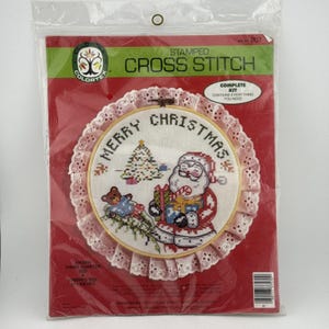 May include: A stamped cross stitch kit in a plastic package. The kit features a completed cross-stitch design of Santa Claus with a Christmas tree and the words "Merry Christmas". The finished size is 10 inches.