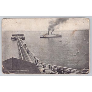 May include: A vintage black and white postcard showing Ericsson Pier in Betterton, MD. A long wooden pier extends into the water with people walking on it. A large steamship with smoke rising from its stacks is in the distance, near small boats.