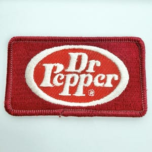 May include: A rectangular embroidered patch featuring a deep red background with a central white oval border. Inside the oval, the text Dr Pepper is stitched in white lettering, accompanied by a small registered trademark symbol in the lower right.