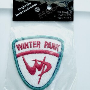 May include: A shield-shaped embroidered patch in a clear plastic bag labeled Imports International. The patch features a white background with a light blue border, pink text reading WINTER PARK, and a stylized pink WP logo on the lower section.