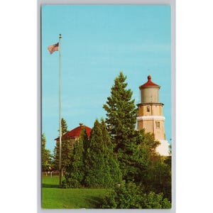 May include: A vintage postcard featuring a tall lighthouse with a red roof and a light-colored tower. Beside it stands a building with a red roof, surrounded by green pine trees. A tall flagpole displays an American flag against a clear blue sky.