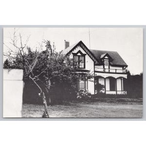 May include: Black and white photograph of a two-story house with a dark roof and white siding. The house has a porch and is surrounded by trees and a grassy yard. The image is a vintage postcard.
