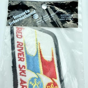 May include: A rectangular embroidered patch in a clear plastic bag labeled Imports International. The patch features a white background with red text reading RED RIVER SKI AREA, accented by blue and red mountain shapes and a yellow snowflake icon.