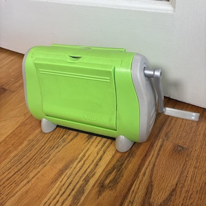May include: A lime green and grey Cuttlebug die-cutting and embossing machine. The machine has a hand crank on the side and is sitting on a wooden surface. The word "Cuttlebug" is printed on the front.
