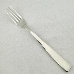 May include: A polished silver-colored metal dinner fork featuring four slender tines and a smooth, flat handle that tapers slightly toward the neck. The utensil has a reflective, metallic finish and a clean, simple design with no decorative patterns.
