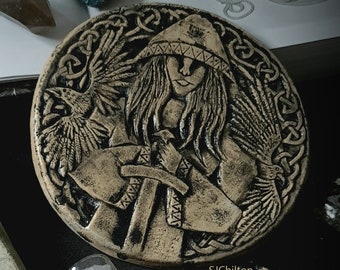 May include: A ceramic round plaque with a raised design of a woman warrior holding a sword and a raven. The woman is wearing a hooded cloak and has long hair. The plaque is surrounded by a Celtic knot design.
