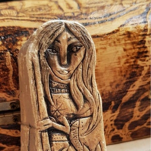 May include: A small, hand-painted ceramic figurine of a woman with long hair, wearing a patterned dress. The figurine is brown and has a textured surface. The figurine is sitting on a wooden surface with a purple crystal next to it. The text "SJChilton" is visible on the crystal.