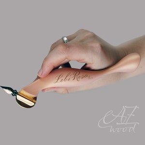 Könnte beinhalten: Ein hellbrauner Kalligraphie-Stift aus Holz, gehalten in einer Hand. Der Stift hat eine goldfarbene Feder und ein geschwungenes Design. Der Text "Lebe Rosa" ist auf den Stift graviert. Das Bild enthält auch den Text "AZ wood".