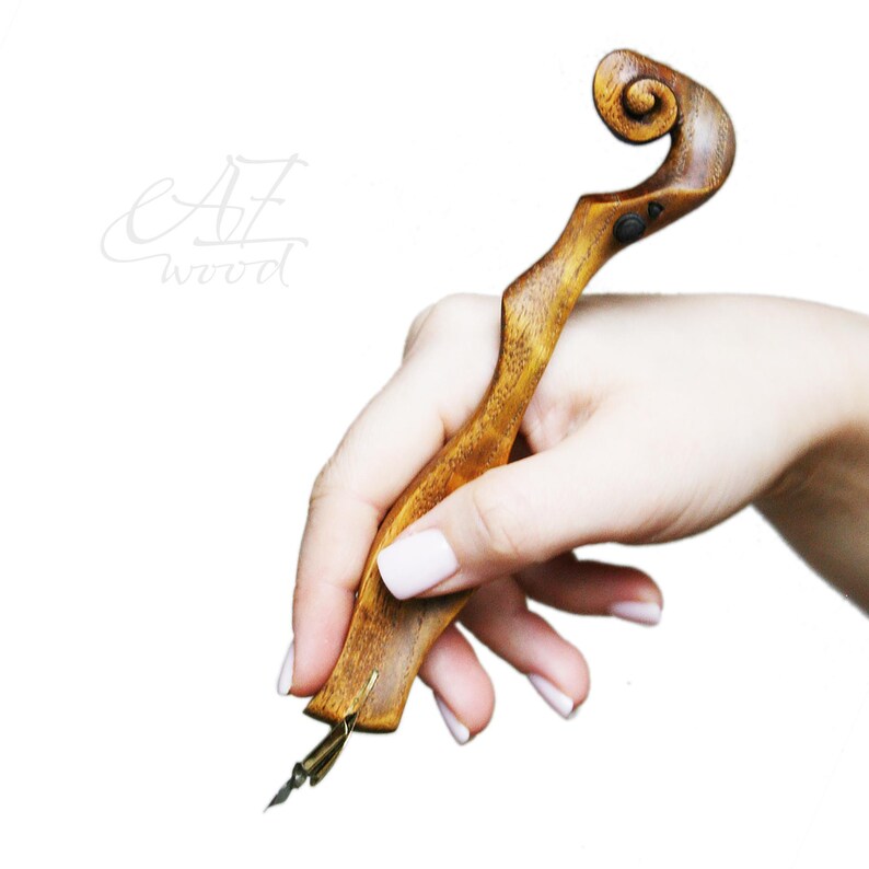 Puede incluir: Una pluma de caligraf&iacute;a de madera con un mango en forma de rollo y una punta dorada. La pluma se sostiene en una mano.