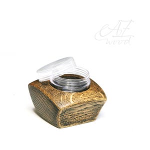 May include: A small, clear plastic jar with a lid sits inside a dark brown wooden container. The container has a square shape with rounded edges. The container is made of wood with a natural grain pattern.