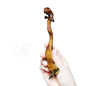 Puede incluir: Una pluma de caligraf&iacute;a de madera con un mango curvo &uacute;nico y una punta de oro. La pluma se sostiene en una mano, mostrando su tama&ntilde;o y forma.