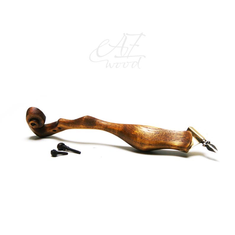Puede incluir: Una pluma de caligraf&iacute;a de madera con un mango de madera marr&oacute;n y una punta de metal. La pluma est&aacute; apoyada sobre una superficie blanca. El mango es curvo y tiene un dise&ntilde;o en espiral en la parte superior. Hay dos peque&ntilde;as piezas de metal negro en la superficie junto a la pluma.