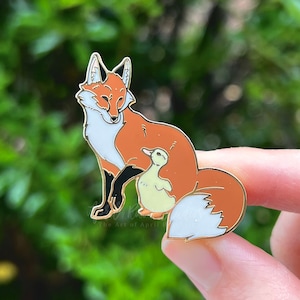 May include: A gold-tone enamel pin featuring a red fox with a white chest and a yellow duckling nestled in its fur. The pin is set against a blurred green background.