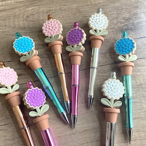 May include: A set of eight flower-shaped pens with different colored flowers and metallic finishes. The pens have a flower design with a pot and stem.