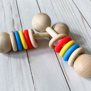 May include: Wooden baby rattle with colorful silicone rings. The rattle has two wooden balls on either end and a wooden handle in the middle. The silicone rings are red, yellow, blue, and white.