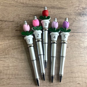 May include: Five silver pens with flower toppers. The flowers are in different colors: red, pink, light pink, purple, and lavender. Each pen has a white pot with a number on it: 1, 2, 3, 4, and 5.